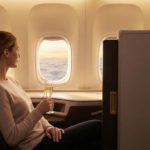 A passenger in a private business-class suite enjoys a glass of champagne while looking out the window at a sunset, emphasizing a serene journey.